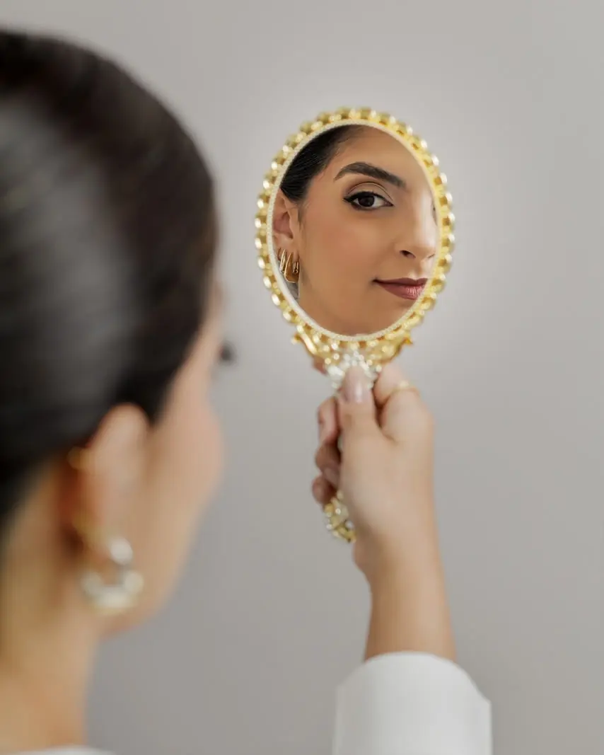 Patrícia de costas, segurando um espelho oval dourado que reflete o rosto dela com sobrancelha e maquiagem naturais