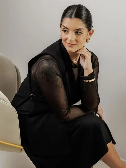 Patrícia Almeida de roupa preta, sentada em uma poltrona clara, olhando para o lado