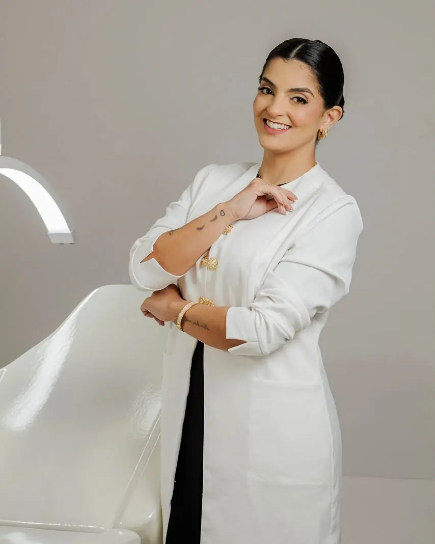 Patrícia de jaleco branco, sorrindo, em pé ao lado da maca do estúdio
