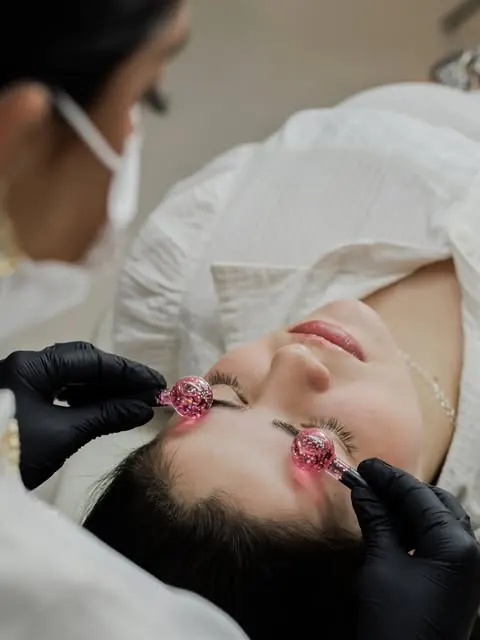 Massagem com esferas geladas rosadas sobre as sobrancelhas de uma cliente deitada, feita com luvas pretas